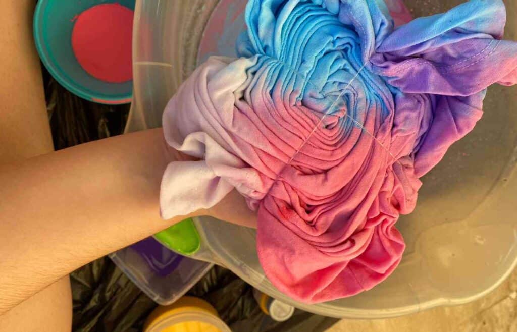 An arm is holding a vibrant, freshly dyed tie-dye shirt over a bucket of water, preparing to rinse it. 