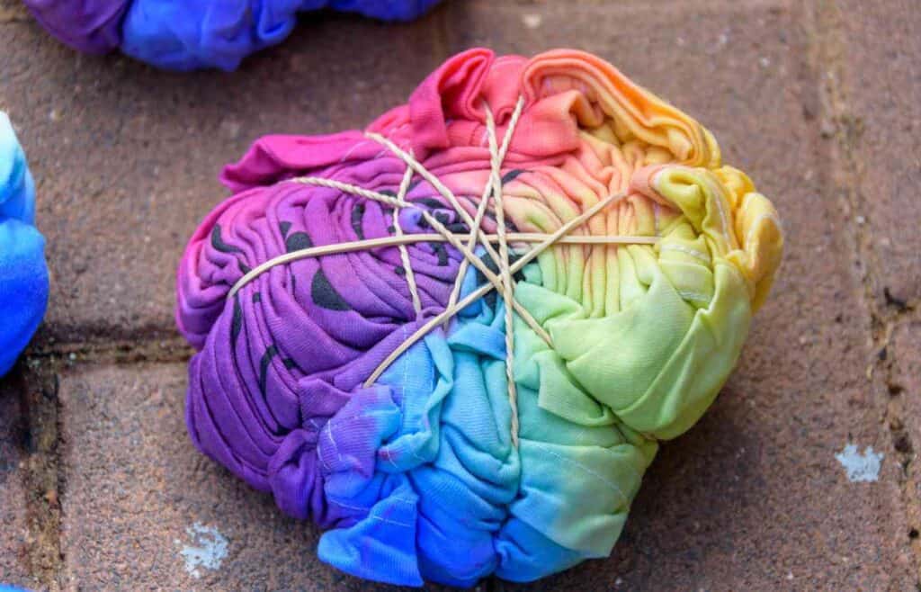 A tie-dye shirt bundled tightly with twine, preparing to dry. The fabric is gathered into sections, held in place by several loops of twine, creating distinct folds.