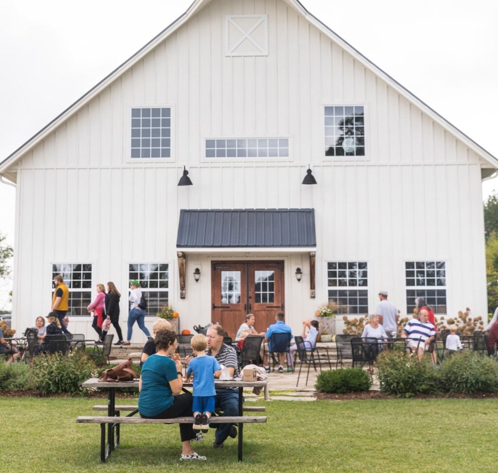 Visitors enjoying outdoor seating in front of the white barn at Peck & Bushel Organic Fruit Company in Colgate, with families gathered for apple picking and farm activities.