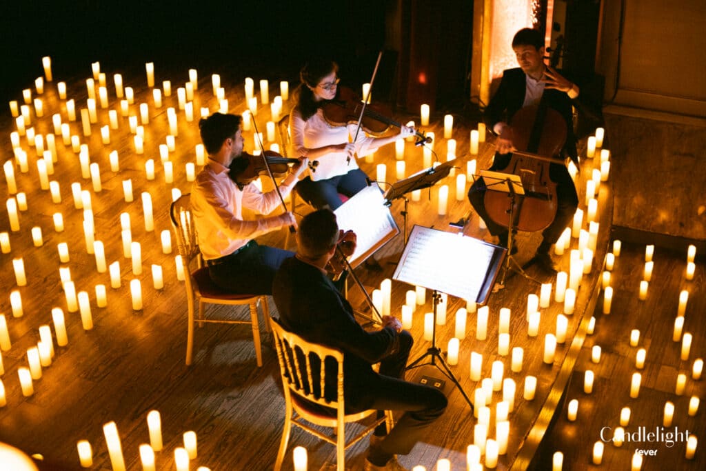 A string quartet performs in a dark room illuminated by numerous candles on the floor, creating a warm, magical ambiance.