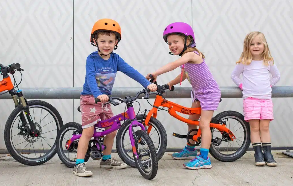 Three kids smile looking at the camera while sitting on their glider bikes in bright colored helmets.