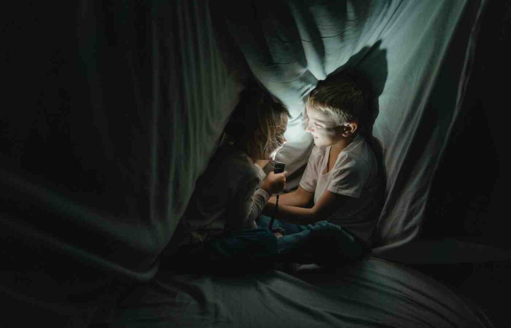 Two children sitting inside a makeshift tent, playing with a flashlight in the dark. The soft light illuminates their faces, creating a cozy atmosphere.