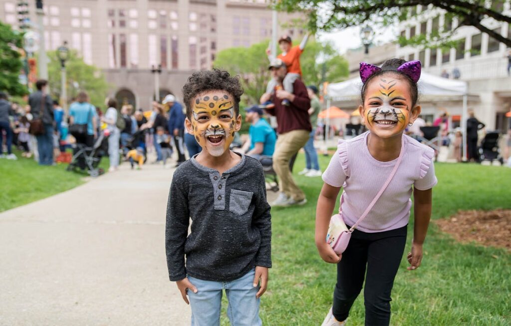 Two children with face paint smiling at Jack-O-Lantern Jubilee in Red Arrow Park, Milwaukee, surrounded by fall festival activities and families enjoying the event.