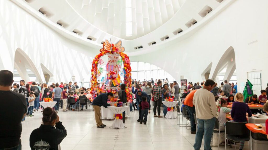 Families and visitors participating in Day of the Dead celebrations at the Milwaukee Art Museum, with a colorful altar as the focal point and activity tables in the background.