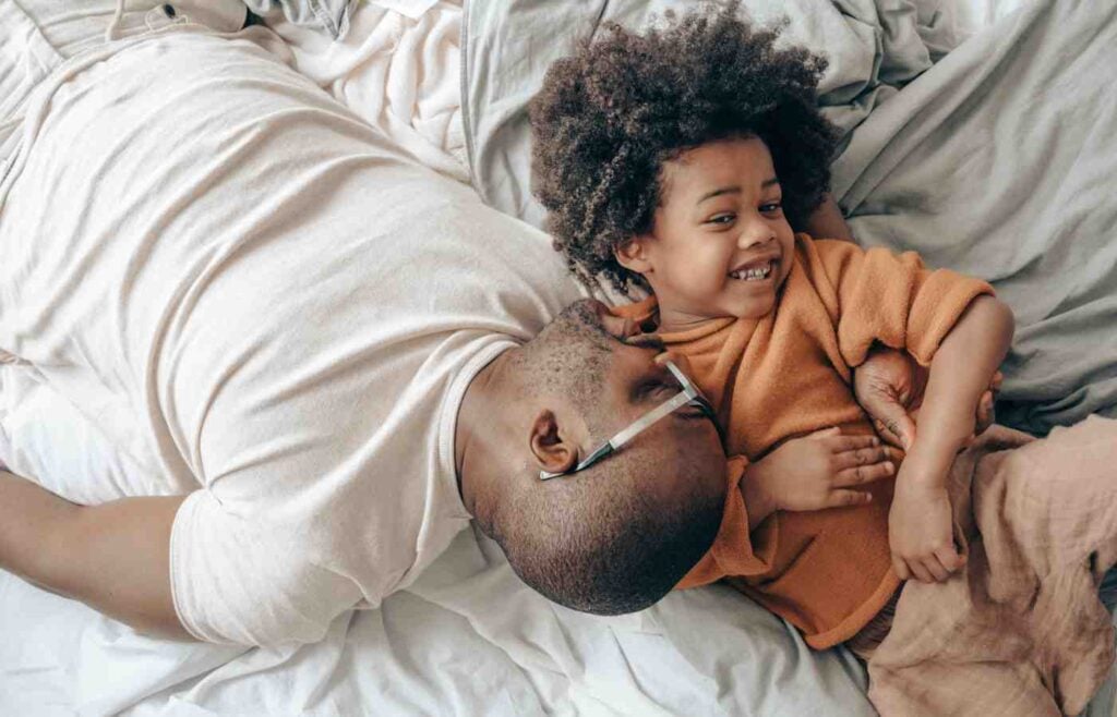 A father playfully lying on the bed with his young child, both smiling and enjoying a lighthearted moment together.