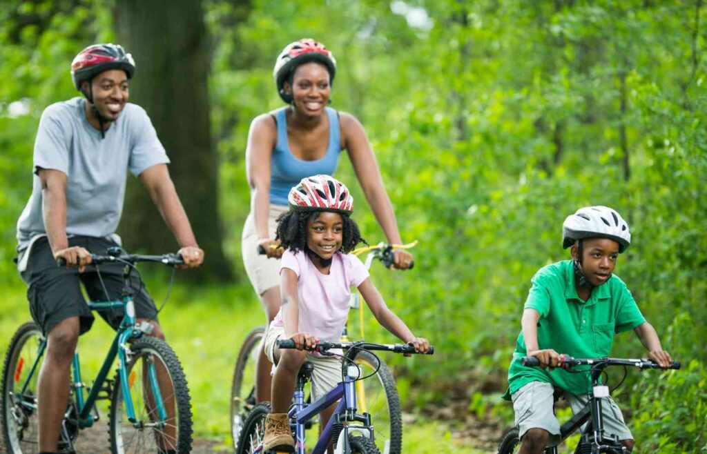 Family with two young kids and a mom and a dad riding through a bike path through a lush green forest on their bikes smiling with helmets on.