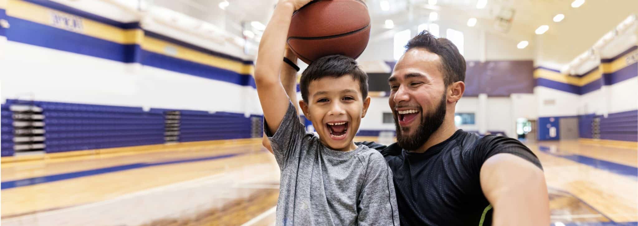 The mid adult father laughs and takes a selfie while his son holds a basketball on his head.