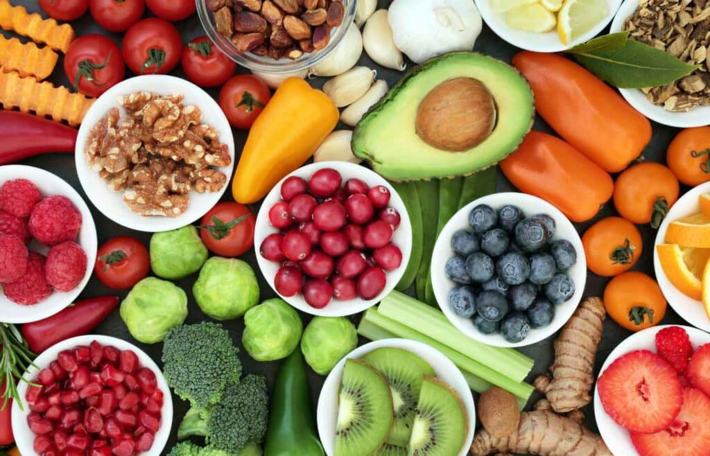 A vibrant assortment of healthy foods including fresh fruits, vegetables, nuts, and seeds. Bowls of blueberries, raspberries, walnuts, and cranberries are surrounded by whole avocados, tomatoes, bell peppers, celery, broccoli, and orange slices, emphasizing colorful, nutrient-rich ingredients.