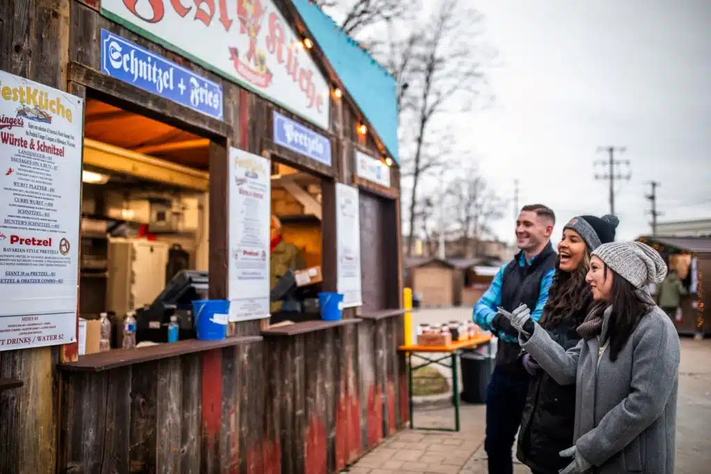 Visitors smiling and ordering food at a schnitzel and pretzel stand at the Wauwatosa Christkindlmarkt, with a festive market atmosphere in the background.