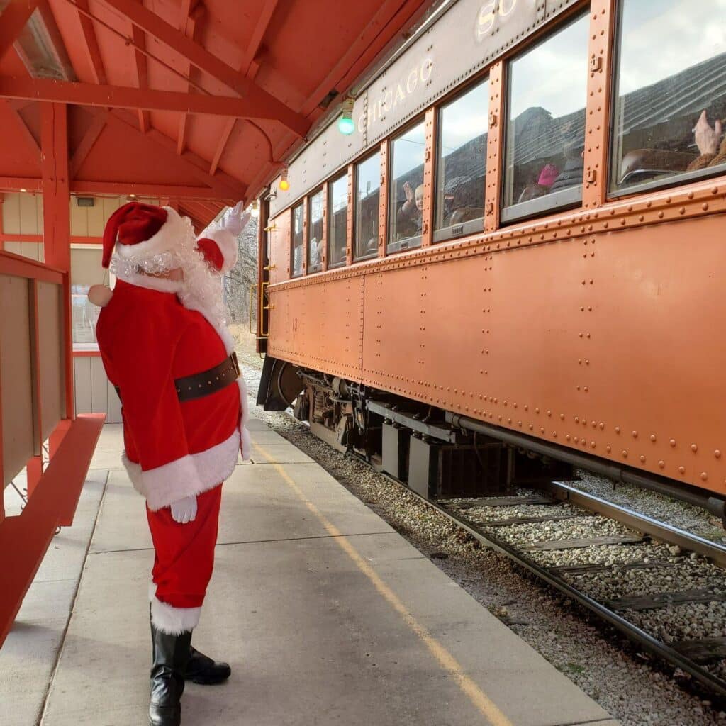 Santa Claus at the platform of the East Troy Depot, waving at passengers on a vintage train decorated for the Christmas season. Santa, dressed in his iconic red suit, is welcoming families as they prepare to embark on a magical holiday journey.