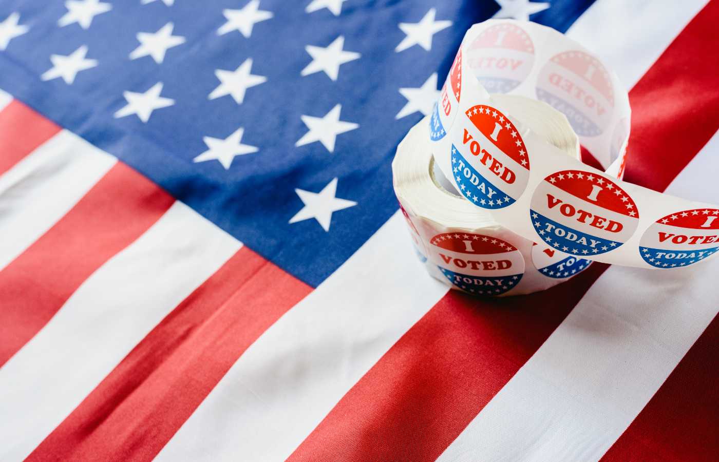 American Flag laying on a table with a roll of stickers that say I Voted with red, white, and blue colors. 