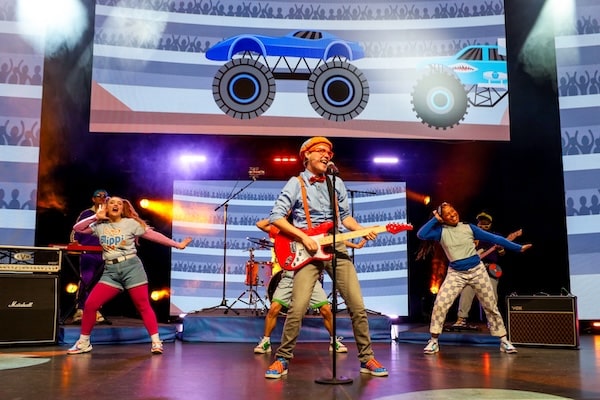 Blippi and friends performing on stage with high energy in the Blippi: Join the Band Tour. Blippi, wearing his signature orange suspenders and hat, rocks out on a red electric guitar while dancers and musicians join him for a lively performance. A colorful monster truck graphic is displayed on a large screen behind them, adding to the excitement for young fans.