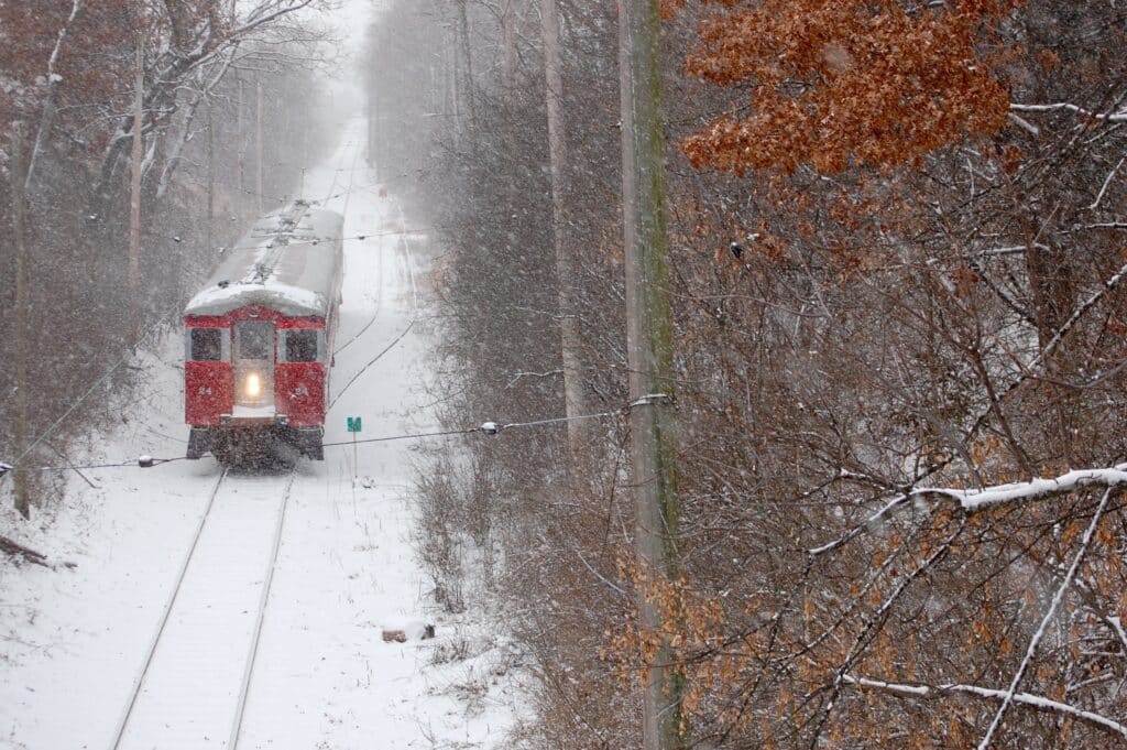 
This image shows a vintage red train moving through a snowy landscape, surrounded by leafless trees and a serene winter setting. The snow is falling gently, covering the tracks and the ground, creating a peaceful, festive atmosphere. The train, with its warm, glowing headlight, looks inviting as it makes its way through the woods, evoking the magic of the holiday season. This is likely the Christmas Train on its journey to Santa’s Workshop, a beloved family-friendly tradition at the East Troy Railroad Museum.