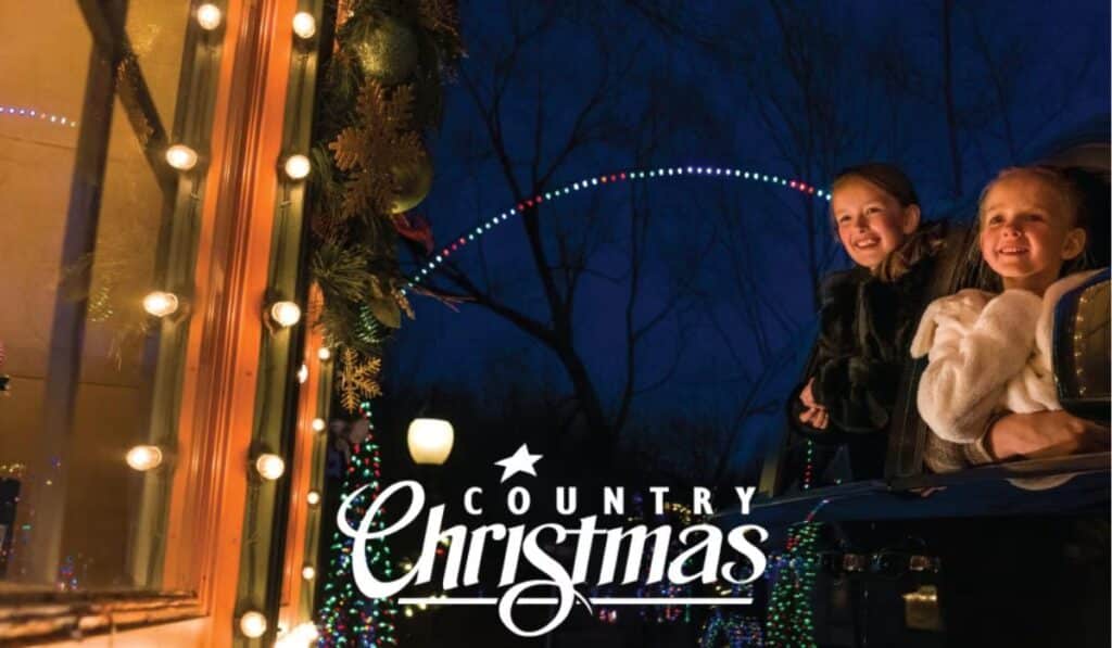 Two smiling children, bundled in warm clothing, lean out of a car window to enjoy the dazzling light displays at Country Christmas. The festive holiday lights and decorations create a magical nighttime setting.