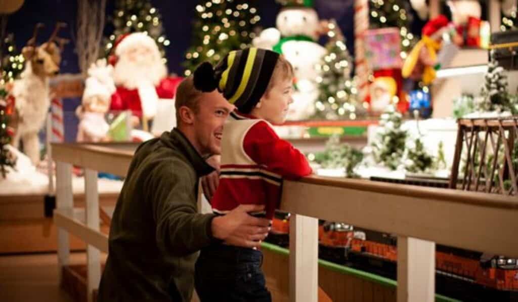 A father and young child look on with excitement at a model train display inside Christmas Village, surrounded by twinkling lights, decorated trees, and holiday figurines.