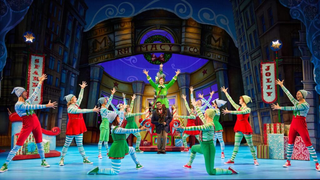 A lively scene from Elf the Musical with actors dressed as elves, dancing around a colorful Christmas-themed set.