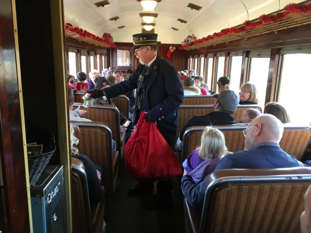 festive interior of the train, where families are seated and enjoying the ride. A conductor dressed in traditional uniform hands out small gifts to passengers, adding to the joyful atmosphere. The train is adorned with holiday decorations, creating a warm, nostalgic feel perfect for the Christmas Train experience.