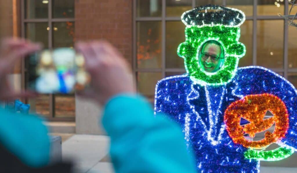 A person stands with their face peeking through a festive Frankenstein cutout made of bright green, blue, and orange lights at Halloween Village in Baird Commons, Milwaukee, holding a pumpkin light decoration. Another person takes a photo, capturing the Halloween spirit.