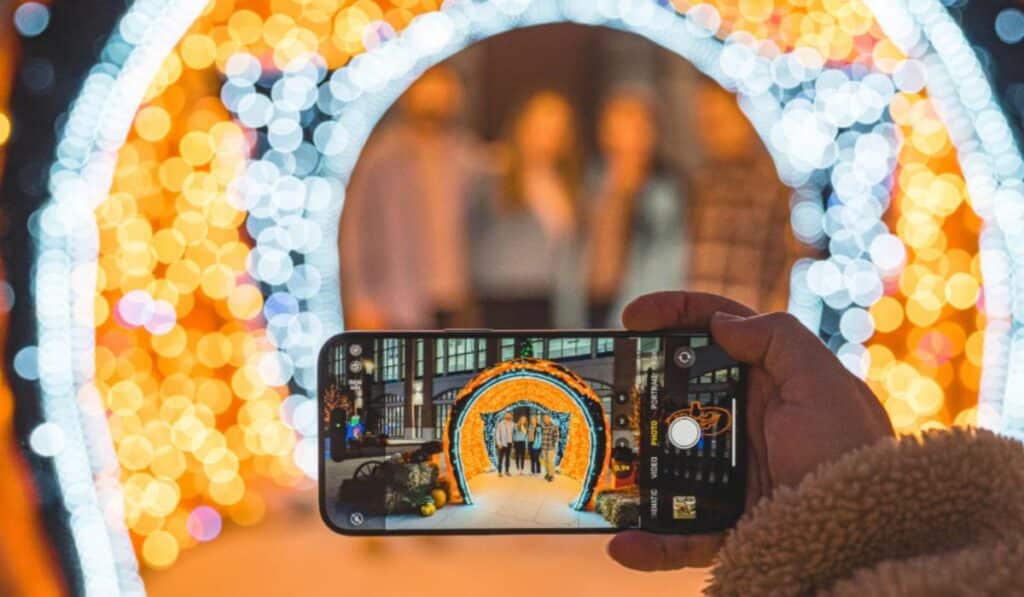 A person holds up a smartphone to capture a family photo through a large, illuminated archway made of orange and white holiday lights at Halloween Village in Baird Commons, Milwaukee. The background features a blurred display of festive lights, creating a magical, glowing atmosphere.