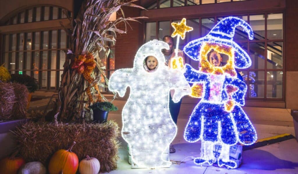 Two children stand behind light-up figures of a ghost and a witch at the Halloween Village in Baird Commons, Milwaukee. The scene is decorated with pumpkins, hay bales, and a corn stalk, giving a festive fall and Halloween feel.