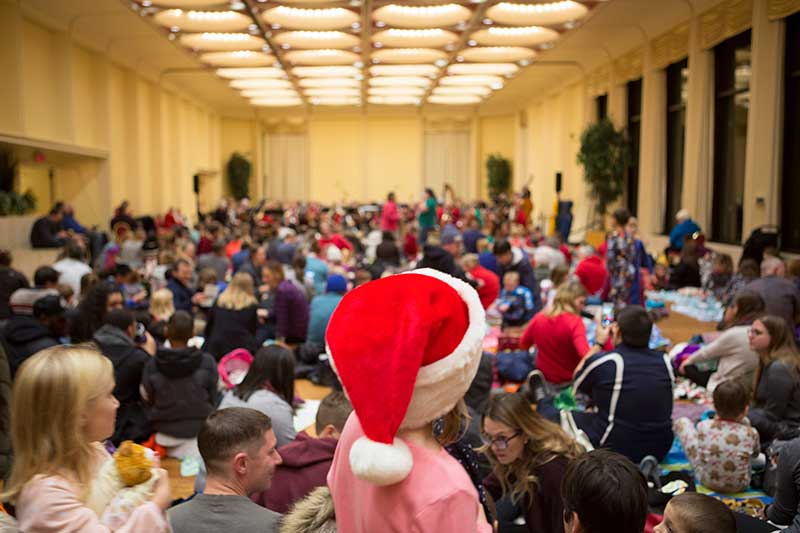 A crowd of families, some wearing Santa hats, sit on the floor and chairs at a Holiday Pajama Jamboree concert, creating a casual and cozy atmosphere.