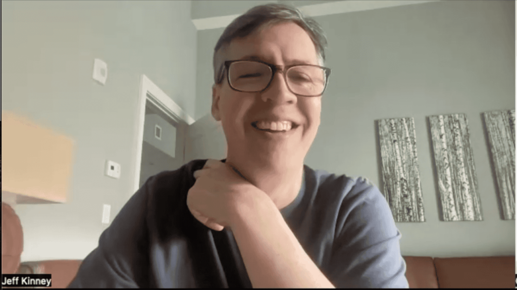 Jeff Kinney smiling during a Zoom interview, wearing glasses and a casual shirt, seated in a warmly lit room with abstract artwork on the wall in the background.