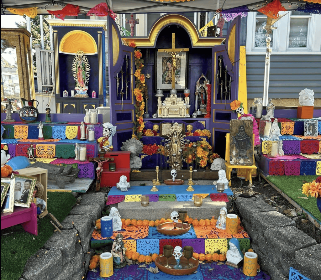 A vibrant outdoor Day of the Dead altar decorated with colorful papel picado banners, marigold flowers, candles, and religious statues. The ofrenda features multiple tiers with framed photos, skulls, and various personal items representing loved ones.