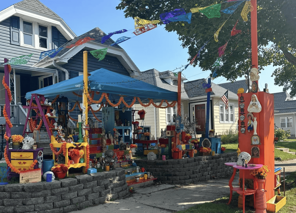 A wide view of a large, multi-tiered community Day of the Dead ofrenda set up in front of a house. The altar is adorned with papel picado, bright marigold garlands, religious figures, and photos, with a blue canopy covering part of the display.