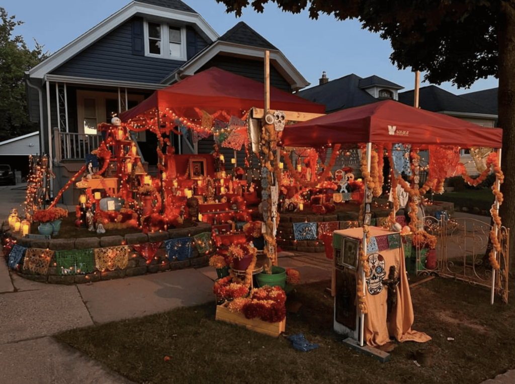 A warmly lit Day of the Dead altar at night, illuminated with orange lights and candles. The scene features multiple tiers of offerings including marigold flowers, skulls, and photos, creating a glowing tribute to loved ones.