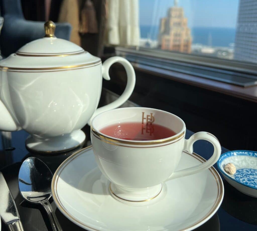 A cup of vibrant red tea in an elegant white teacup with a matching teapot, set against a cityscape view from The Pfister Hotel.