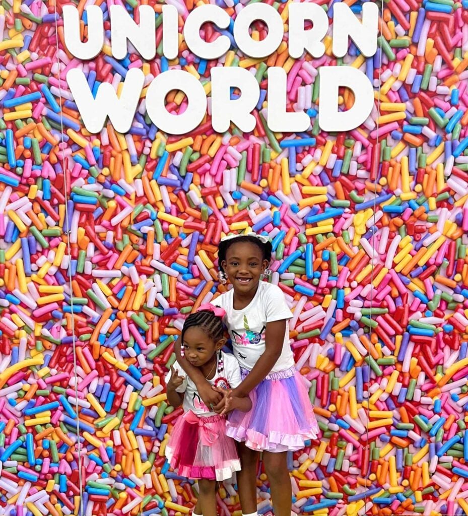 Two young girls posing in front of a vibrant backdrop filled with colorful sprinkle-like decorations, under the sign 'Unicorn World.