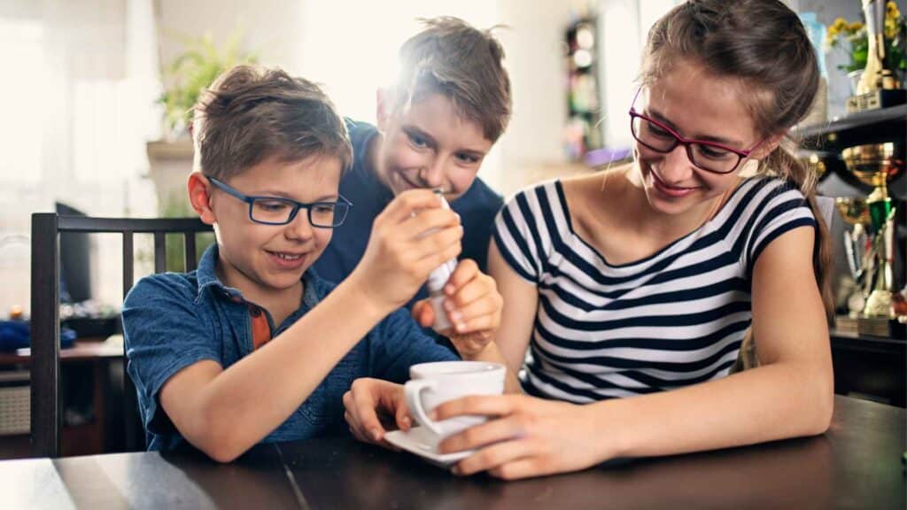 Three kids sitting at a kitchen table putting salt into their parent's coffee cup.