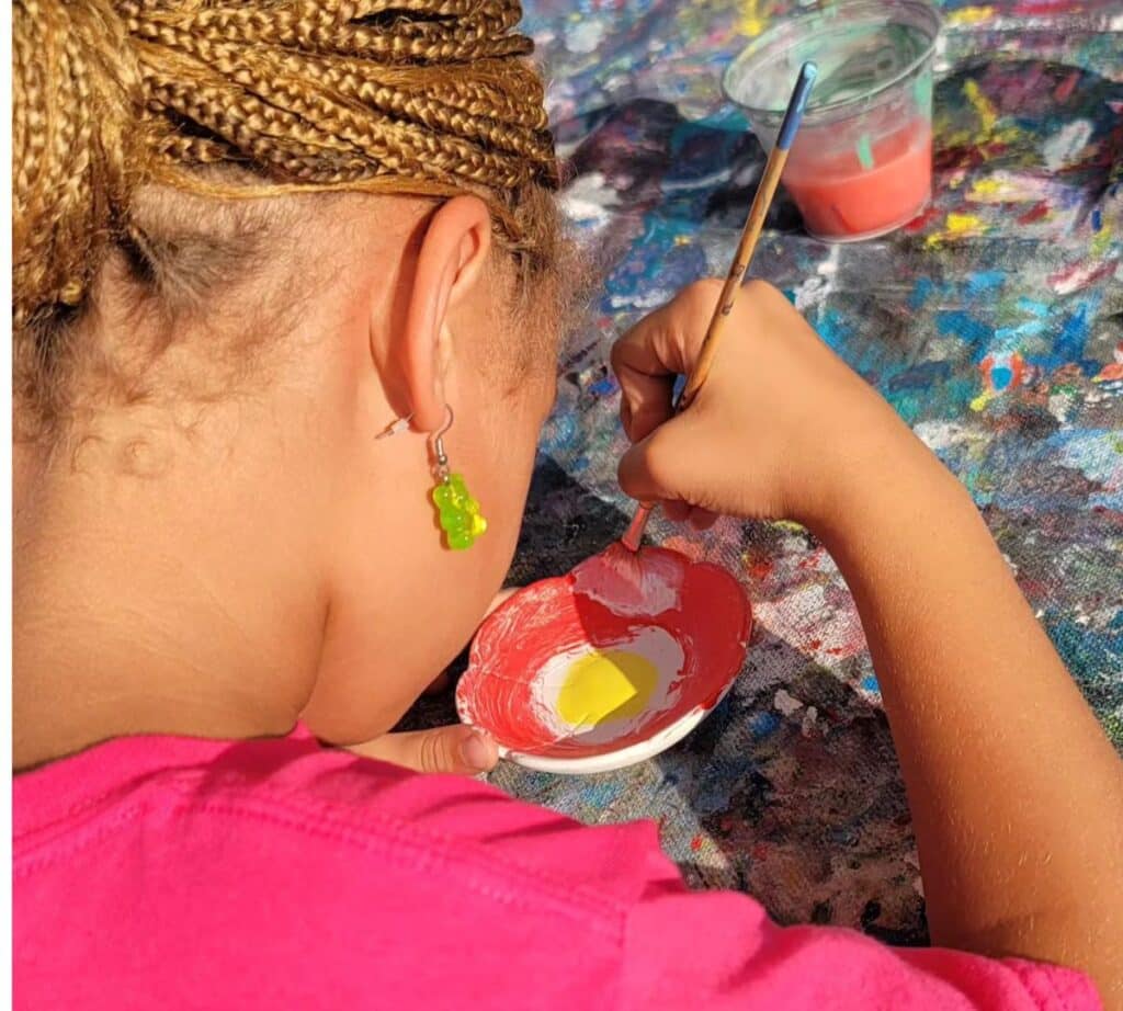 A young artist carefully painting a small ceramic bowl at Vibez Creative Art Studio. The artist is wearing a bright pink shirt and has braided hair with green gummy bear earrings. The table is covered in a colorful, paint-splattered cloth, with a cup of water and paintbrush nearby, creating a vibrant and creative atmosphere.