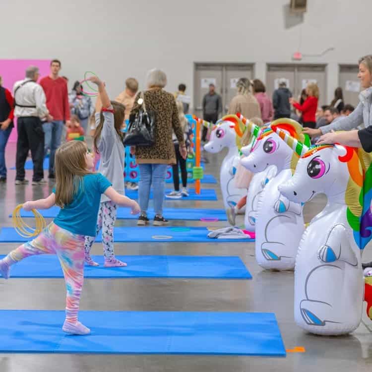 Children playing a ring toss game with inflatable unicorns at an indoor event, surrounded by families and colorful mats.