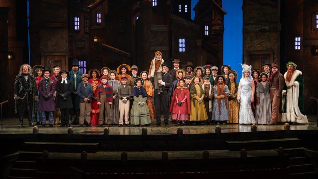 The full cast of A Christmas Carol takes a group photo on stage, dressed in period costumes with a classic Dickensian backdrop.