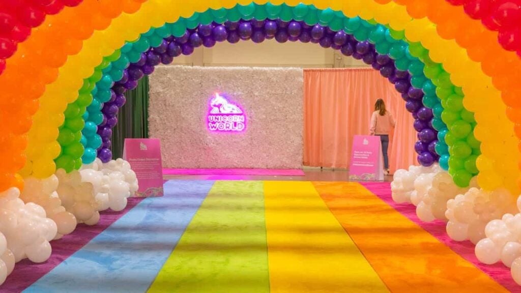 Rainbow archway made of balloons leading to a bright pink sign reading 'Unicorn World' at an indoor event space.