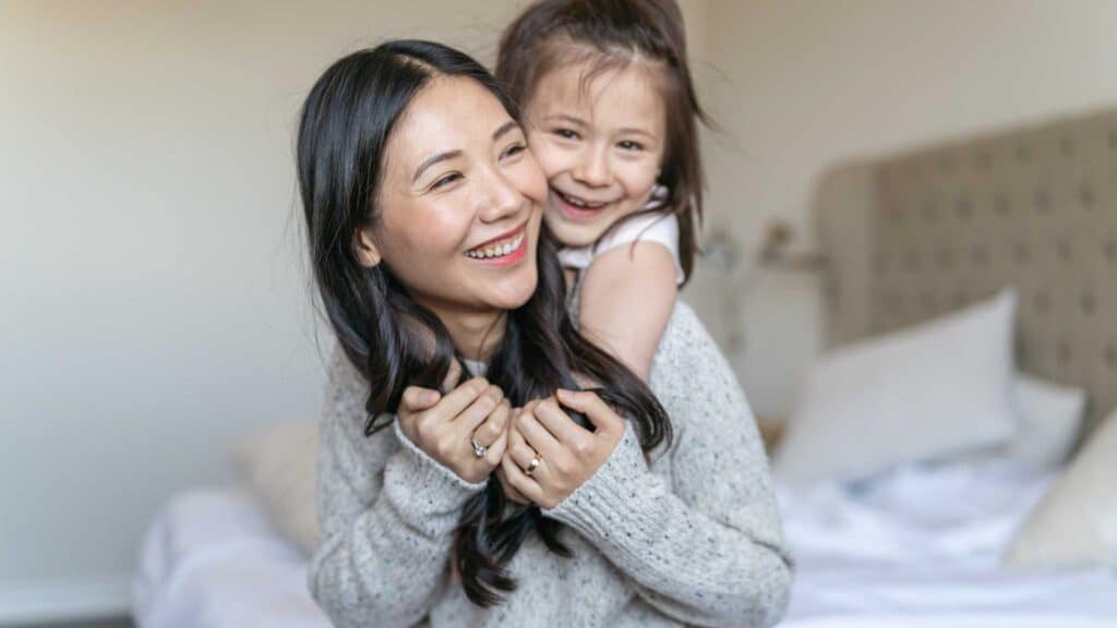 A young daughter piggy backed onto her mom with her arms wrapped around her shoulders in a bedroom with an unmade bed. Both are smiling and enjoying each other.