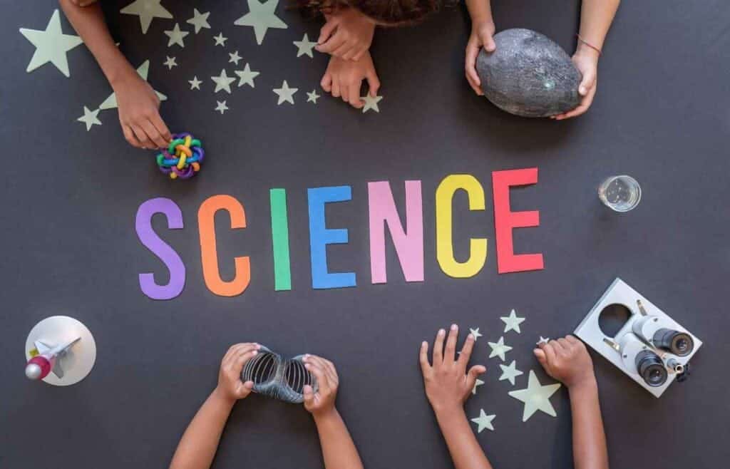 Children’s hands holding various science objects, including a globe and slinky, around colorful letters spelling 'SCIENCE' on a black background with stars.