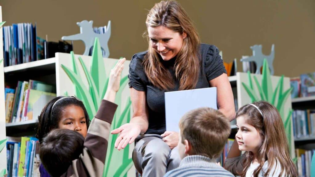 Students listening to story time in a library with their teacher, one is raising their hand.