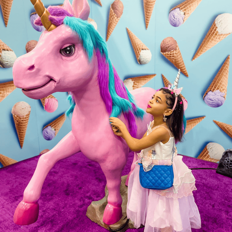 A young girl in a unicorn-themed outfit standing beside a large pink unicorn statue with ice cream cone decorations on the wall behind her.